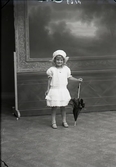 Ateljébild. Barnporträtt. Flicka stående i helfigur i ljus sommarklänning och mössa, hållande ett solparasoll i vänster hand. Beställare: Frk. Reinholdsson.