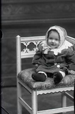 Ateljébild. Babyporträtt, baby sittande i helfigur i ytterkläder med spetskrage och spets även på mössan. Beställare: Fru Johansson, Väderkvarnen, Falkenberg.