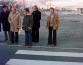 En samling män står vid övergångsställe vid Frölundagatan i Åby, Mölndal, omkring 1975-1980. I bakgrunden ses idrottshuset.