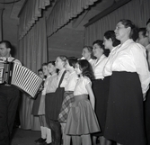 Bild tagen i samband med flyktingar ifrån Ungern 1956. Folk som står på scen och en man som spelar dragspel, förmodligen i Godtemplargården. Hösten 56 - våren 57.