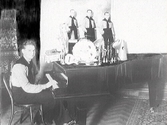 En man sitter vid en flygel, troligen i matsalen på Varbergs Hotell. På flygelns lock står en miniatyrmodell av okänd orkester med tre män och instrument, likadant klädda som pianisten.