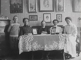 Interiör. Fyra kvinnor sitter i en soffa vid ett dukförsett bord, där två inramade fotografier står. Vid bordets vänstra sida står en pojke. På väggen bakom dem hänger flera tavlor och fler fotografier.