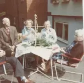 Fem personer sitter runt ett bord utomhus, Brattåshemmet vid midsommar, 1970 - 80-tal. På bordet står en obunden midsommarstång samt en hög med gröna blad. Från vänster: Okänd person som ses delvis. 2. Anselm Zackrisson. 3-5. Namnuppgifter saknas. Relaterat motiv: A4274.