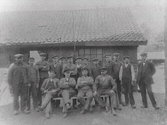 Arbetsstyrkan på Varbergs Mekaniska Verkstad år 1916. Männen samlade i olika typer av arbetskläder utanför en av byggnaderna. Fyra av dem sitter på en bänk framför övriga.
