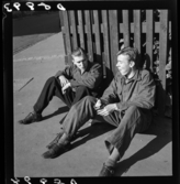 539 Söder. Två unga män sitter på gatan lutande mot ett staket.