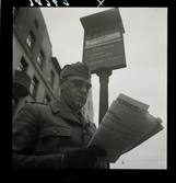 558 Wilhelm Moberg. Vilhelm Moberg står i uniform och läser tidningen vid en spårvagnshållplats.