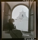 568 F. Stieg Trenter, Torremolinos. En kvinna sitter i ett öppet fönster. Ett klocktorn syns på andra sidan gatan.
