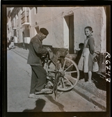 568 F. Stieg Trenter, Torremolinos. En man slipar en yxa ute på gatan. En flicka tittar på.