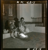 568 F. Stieg Trenter, Torremolinos. En flicka och en kvinna sitter på tröskeln utanför ett hus. En eld brinner på ett fat framför dem.