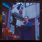 585/8 Facit Tokyo Daberg. En man telefonerar och tittar på sitt armbandsur, vid en station med flera röda telefonapparater.