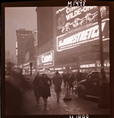 1690 New York, allmänt. Gatubild nattetid. Neonljus, reklam. Två kvinnor kommer gåendes under ett paraply.