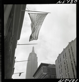 1690 New York allmänt (N.Y. Herald Tribune). Gatubild. Amerikansk flagga hänger från fasad, Empire state buildning i bakgrunden.