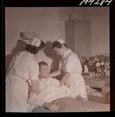 1714 En sjuksköterskas arbetsdag på södersjukhuset. Ett par sköterskor hjälper en patient i en sjukhussäng.