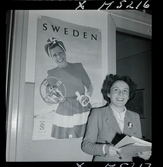 1717/K Istanbul allmänt. En leende kvinna med papper i händerna står framför en affisch som gör reklam för Sweden.