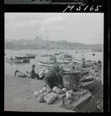 1717/K Istanbul allmänt. Gatuförsäljare. En man säljer frukter på kajen. Roddbåtar på vattnet.