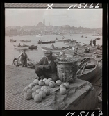 1717/L Istanbul allmänt. En man sitter på kajen och säljer frukt, bakom honom roddbåtar i vattnet.