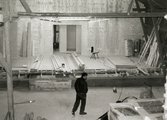 Nya Teaterhuset byggs upp 1997 - 1998. En bild inomhus på ett stort rum. På golvet står det en person som tittar åt höger, bilden är tagen bakifrån. Framför personen från markhöjd är det en uppbyggnad som det ligger lösa plankor på. På uppbyggnaden står det även en stol, och inplastade förpackningar med isolering och en lång stege står lutad mot väggen. Det är en stor öppning i mitten av väggen in till nästa rum. Man ser ladans bjälkar rama in fotot.