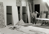 Nya Teaterhuset byggs upp 1997 - 1998. En bild inomhus på ett rum med träskivor som väggar. En person sågar i en stor träskiva som ligger på en sågbock. På golvet ligger det plankor i mindre storlekar och en klump med sladdar och grenuttag. Mot väggarna står det lutade olika storlekar av träskivor.
