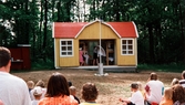 Sommarspel/barnteaterföreställning, troligtvis 1999. Vid Byxorna i Gunneboskogen (korsningen Christina Halls väg/John Halls väg). Några barn agerar i en liten gul byggnad. Framför dem sitter publik och ser på.