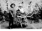 Två små barn utomhus där det äldre står bredvid en baby som sitter i en ålderdomlig anordning av kombinerad barnstol och kärra. Babyn är nedtill insvept i en filt och pojken bär en tjock stickad rock och mössa. Bakom dem  syns en rabatt längs en tegelvägg. Fotograf okänd. 1910-tal. (Se även bildnr C664)