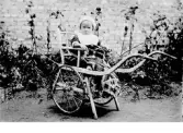 Litet barn som sitter i en ålderdomlig anordning av kombinerad barnstol och kärra, som placerats utomhus intill en rabatt längs en tegelvägg. Barnet är nedtill insvept i en filt. Fotograf okänd. 1910-tal. (Se även bildnr C665)