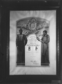 Fotografi av diplom: Industri- och slöjdutställningen i Gefle 1901.
Diplom för silvermedalj. Två män på var sida om textfältet. mannen till vänster är en arbetare med slägga i hand. Mannen till höger håller och läser en stor bok.