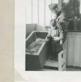 Karl Gustaf Pettersson står bredvid sonen Lennart och tittar ner i en babysäng där det ligger ett litet barn, Backen eller Högen (övre gården) 1960-tal.