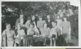 Fem personer sitter vid ett bord och fikar. Bakom dem står åtta personer, Alegårdsslätt 1940-tal. Sittande vid bordet, från vänster: 1. Svea Johansson. 2. Anna Johansson. 3. Stina Johansson. 4. Tora Frej Johansson. 5. Ivar Johansson. Bakre rad, från vänster: 1. Rolf Benkel. 2. Ida Johansson. 3. Maja Johansson Benkel. 4. Gustav Benkel. 5. Kerstin eller Greta Johansson. 6. Okänd. 7. Ingeborg Johansson Andersson. 8. Rose Maj Benkel Ekström.