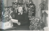 Svea* Gudviva Fredrika Johansson(1892 - 1976) sitter omgiven av blommor. Hon håller ett papper i handen, Heljered Västergård 