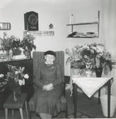 Uppvaktning av Hulda Eriksson (1874 - 1960) år 1959 på Brattåshemmet där hon bodde på 1950-talet. Hon sitter finklädd i en fåtölj med en huvudbonad på huvudet. Rummet är fyllt av blommor. Uppväxt på Heljered Västergård 3:3 