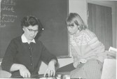 Handarbetslärarinnan Rut Fonell (1903 - 2005) undervisar eleven Kristina Olsson, Brattåsskolan 1965. I bakgrunden skymtar 