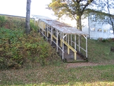 En sluttande fristående trappa med tak, sedd från vänster, där människor kan köa till alternativt lämna Hällesåkers dansbana år 2010. Till höger ses en vit byggnad. Området är omgivet av växtlighet samt är inhägnat med staket.
Relaterade motiv: 2024_1119 - 1137.