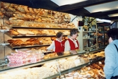 Två kvinnliga personal står bakom en bröddisk med hyllorna fulla av diverse bakverk, K-marknad i Mölndals centrum 1980-tal. Framför bröddisken står en kvinnlig kund.