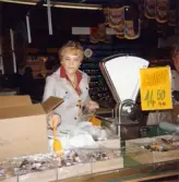 En kvinnlig expedit håller en plastslev i enda handen, med den plockar hon smågodis (lakritskonfekt) och lägger i en plastpåse för att sedan väga det på vågen som står bredvid, Domus vid Nya torget 1970-tal. Vid sidan av vågen står en skylt med texten: GODIS 14.50 1/5 kg. Bortanför den skylten skymtas en annan kvinna.