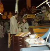 En manlig och en kvinnlig butikspersonal står vid ett bord och samtalar. Båda är klädda i grå-beiga kläder. Bredvid dem på golvet står staplade bruna backar, fyllda med långa, plast-förpackade rulltårtor. Närmast i bild ses en skymd kvinna som erbjuder provsmakning. Hon har gjort små bakelser bestående av en skiva rulltårta toppad med vispad grädde och bär. I taket hänger flertalet skyltar med texten 