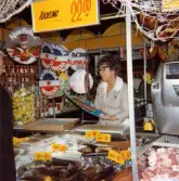 En kvinnlig expedit står bakom fiskdisken och håller en stor inplastad fisk i handen. Hon pratar med en kund. Framför henne finns en kyldisk fylld med fisk- och skaldjursförpackningar. På hyllorna till vänster står staplade Ajvar-burkar och i en glas-skål finns konserverade förpackningar med pressad citron. Till höger ses en våg. I taket hänger runda pappers-skyltar med texterna 