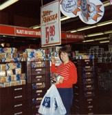 En kvinnlig kund står vid bröd-avdelningen och håller ett paket jättefranska samt på plastpåsar från Coop i handen, Domus Mölndal 1970-tal. På golvet står staplade bruna plastbackar fyllda med bröd. Ovanför backarna finns en skylt med texten 