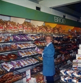 En kvinnlig expedit, klädd i blå rock, står vid en vägg med uppsatta brödhyllor fyllda av olika slags bröd och kaffebröd, Domus Nya Torget 1970-tal. Ovanför hyllorna finns en lång grön skylt med texten 