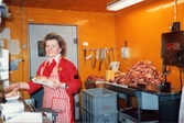 En kvinna står i ett orangefärgat inplastat rum avsett för styckning av kött. I händerna håller hon styckdetaljer. På väggen snett bakom hänger olika typer av knivar och sågar. På en diskbänk bakom henne finns mer styckdetaljer. På golvet står gråa plastbackar, Domus Mölndals Centrum 1970-tal.