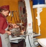 En kvinna står i ett orangefärgat inplastat rum avsett för styckning av kött. Hon står vid en stor skärmaskin och skär ett stort köttstycke i bitar. På väggen i bakgrunden hänger olika typer av knivar och sågar. Närmast till höger ses ett handfat och torkpapper, Domus Mölndals Centrum 1970-tal.