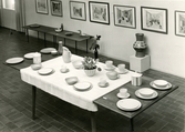 Uppdukat bord med keramik från Bo Fajans, utställning på Gefle Museum 24/4 - 4/5 1956. På väggen hänger tavlor av Josabeth Sjöberg.