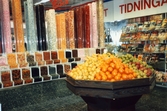 En stor rund disk fylld av färsk frukt, i bakgrunden ses rör fyllda med lösgodis för självplock. I bakgrunden till höger skymtar vägghyllor med tidningar, K-marknad i Mölndals centrum 1990-tal.