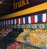 Närbild av en lång disk fylld av frukt och grönsaker som står utmed en vägg, Domus Centrum 1970-tal. Ovanför disken hänger en skylt med texten 