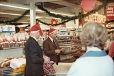 Kjell och Thore har på sig julmössor och delar ut julklappar till kunderna, Domus vid Nya torget julafton 1968. I bakgrunden ses en skylt som hänger i taket med texten 