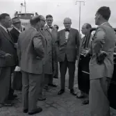 Herrar i kostym. Statsrådet Eliasson bakom bildörren. Ruben Wagnsson står bredvid honomoch tittar in i kameran.