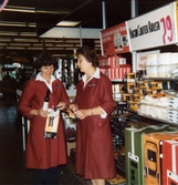 Två kvinnliga expediter, klädda i röda rockar, står i el-avdelningen bland dammsugare, brandvarnare, locktänger och hårfönar, Domus Frölundagatan okänt årtal. En av kvinnorna håller en Hugin Curler Brusch med tillhörande kartong i händerna. Denna säljs för 79:-.