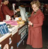En kvinnlig expedit, klädd i röd rock, står bakom en disk och lägger ner varor i en kasse, Domus Frölundagatan okänt årtal. Framför henne står en kvinnlig kund redo att betala. På diskens framsida sitter en lapp med texten FILMINLÄMNING. Nedanför sitter ett vitt klistermärke med den blå texten 