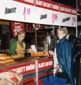 En kvinnlig demonstratris, klädd i grön rock och gult förkläde, står i en bröddisk för provsmakning, Domus Frölundagatan efter 1976. Ovanför bröddisken hänger skyltar med texten 