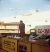 En kvinnlig expedit, iklädd röd rock, står utomhus mitt i en stor gul papperskontainer, Domus Mölndals C okänt årtal. På kontainern står textat AB INDUSTRILEVERANSER RETURPAPPER Tel. 031-51 34 80. I bakgrunden ses en Goman-lastbil samt ett hus (lastkaj) med texten BOKHANDEL. Längre bort skymtar två byggkranar och kyrktornet till Fässbergs kyrka.