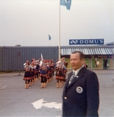 Främst ses en man, iklädd slips och blå kavaj, som står på en parkeringsplats utanför Domus, Nya torget före 1976. Bakom honom står en grupp sameklädda personer och spelar intstrument. I bakgrunden ses Domus som ligger i en blå barack-byggnad.